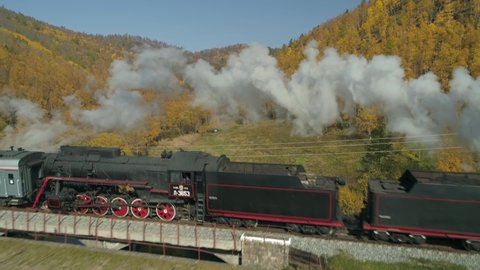 Aerial approach old steam locomotive train rides on scenic ancient stone arched bridge Trans-Siberian Railway railroad. Circum-Baikal lake. Autumn yellow forest shore. White smoke from pipe. Sunny day