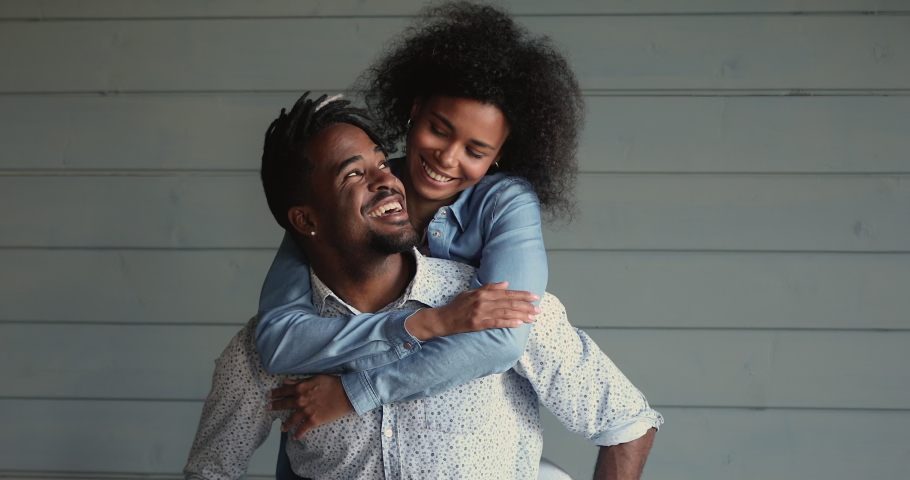 Portrait of candid charming just married African 30s couple in love concept. Happy mixed race wife piggy back beloved husband with dreadlocks wear casual clothes posing on blue wooden wall background | Shutterstock HD Video #1062340777
