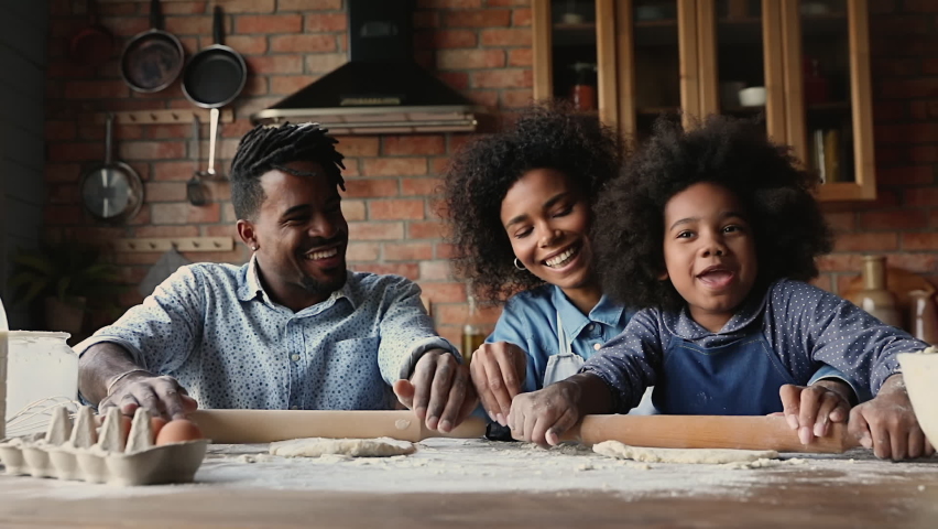 Happy african family with preschool 6s daughter flattening dough using rolling pin cooking together in kitchen. Teach and develop kid, prepare holiday cake for event celebration, common hobby concept Royalty-Free Stock Footage #1062340834