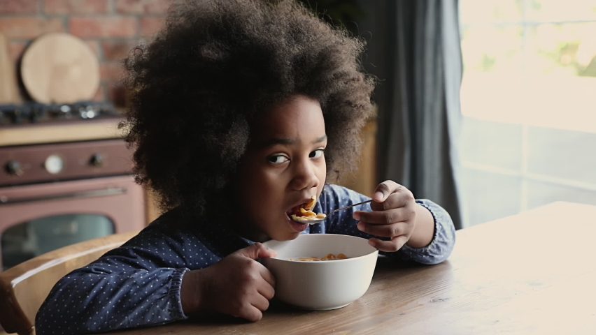 Close up pretty african little girl with curly afro hair sitting at dining table in domestic kitchen eating dry breakfast cereals corn flakes with milk. Balanced meal for child healthy growth concept Royalty-Free Stock Footage #1062340882