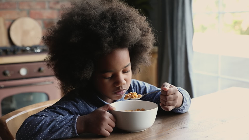 Close up pretty african little girl with curly afro hair sitting at dining table in domestic kitchen eating dry breakfast cereals corn flakes with milk. Balanced meal for child healthy growth concept | Shutterstock HD Video #1062340882