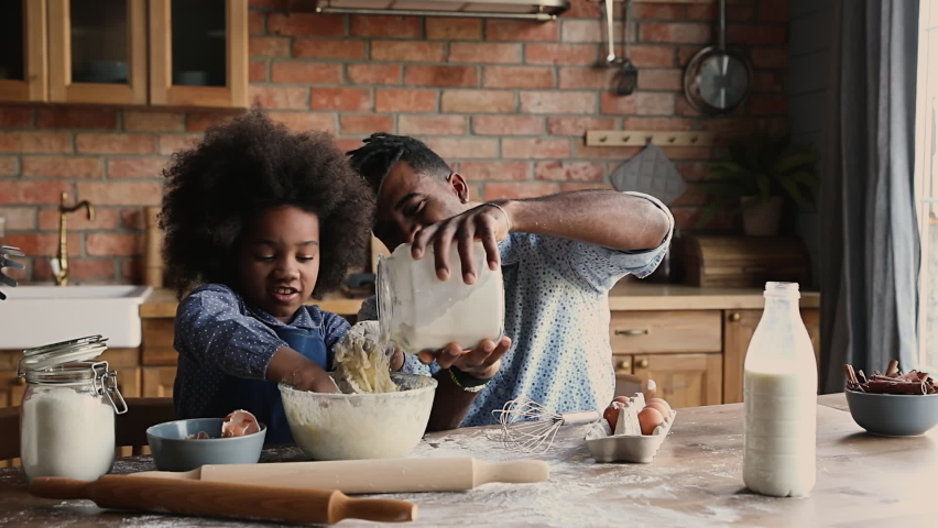 African dad with dreadlocks pour flour while curly haired daughter knead dough in bowl, family prepare surprise sit at dining table in kitchen. Spend time together develop kid, pleasant chores concept Royalty-Free Stock Footage #1062340894