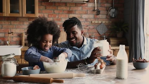African dad with dreadlocks pour flour while curly haired daughter knead dough in bowl, family prepare surprise sit at dining table in kitchen. Spend time together develop kid, pleasant chores concept