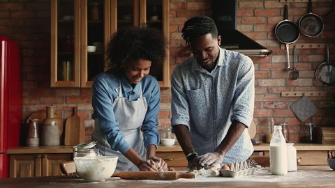 Happy young family African wife and husband standing in cozy kitchen kneading dough cooking together prepare pastries for holiday dinner, celebrate event, enjoy romantic date and communication at home