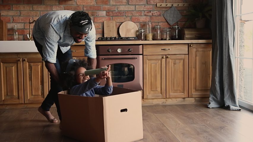 Happy African daddy and kid play pirates in warm kitchen, barefoot dad pushing daughter seated in box hold carton binocular or telescope went looking for treasure. Family having fun at home concept Royalty-Free Stock Footage #1062340936