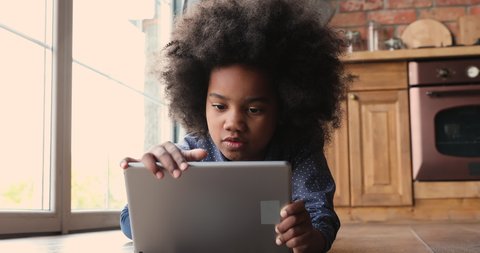 Little african girl lying on warm floor with tablet, look at device screen watch cartoons, listen audio book, close up. Addiction of modern tech usage, parental control, safety, gadget overuse concept
