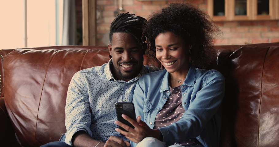 African sport fan couple sit on couch at home watching game match on-line in streaming on smart phone celebrating victory. Happy supporters cheering and exulting after winning on internet bet concept Royalty-Free Stock Footage #1062342772