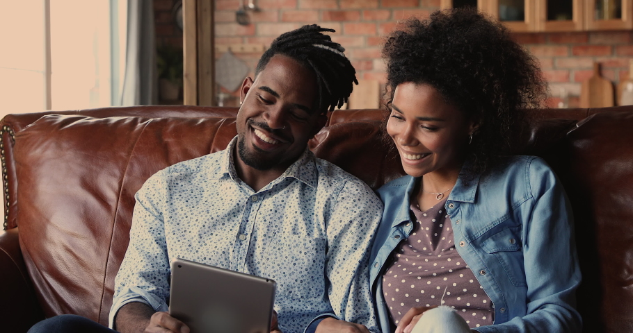 African 30s handsome boyfriend and pretty girlfriend relaxing sit on couch holding tablet, family buy goods for home use e-store on-line e-commerce retail services. Modern tech, new app usage concept Royalty-Free Stock Footage #1062342784