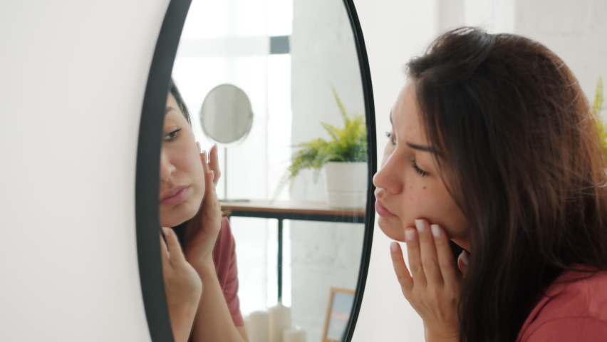 Slow motion of beautiful young woman touching face looking for imperfections on skin in bathroom alone. Modern lifestyle and beauty concept.