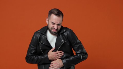 Sick worried displeased young bearded man 20s in black leather jacket posing isolated on orange color background studio. People lifestyle concept. Feeling bad put his hands on stomach abdominal pain