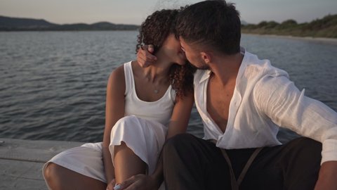 Romance scene of young beautiful heterosexual lovers couple on a pier looking at the sun goes down - Bearded handsome man kiss her Hispanic black curly woman sitting on a wooden old jetty on the water