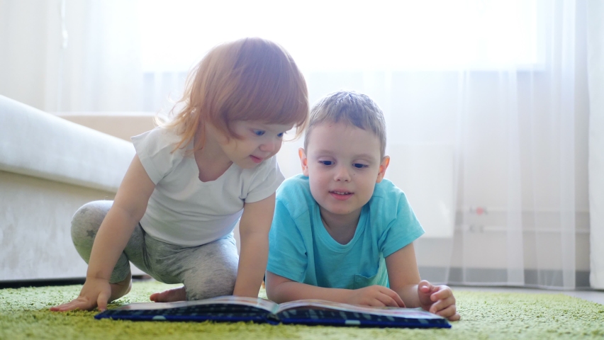 Curious cute preschool redhead girl sitting on carpet floor alone and her brother reading book for her | Shutterstock HD Video #1062350245