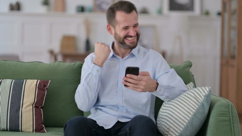 Young Man Celebrating Success on Smartphone on Sofa 