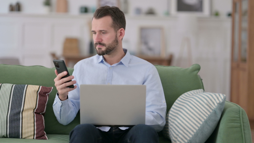 Young Man working on Smartphone and Laptop on Sofa 