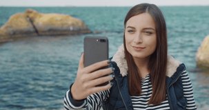 Young beautiful girl waving hand holding smartphone while standing by the sea. Happy female tourist talking on video call from the coast.