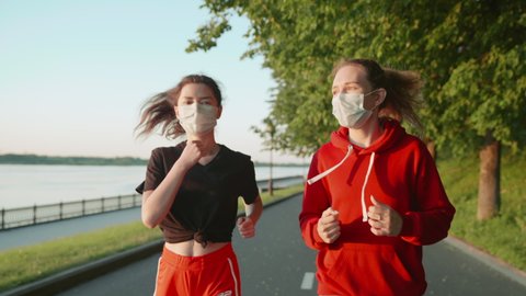 Brunette and blonde run in Covid. New life after quarantine. Girls keep a social distance and run along the embankment in protective masks. Concept of healthy lifestyle, training, jogging.