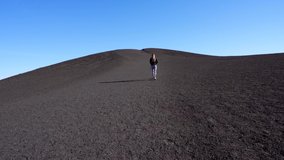 This video shows a lone woman walking on a remote black volcano summit hill with a blue sky behind.