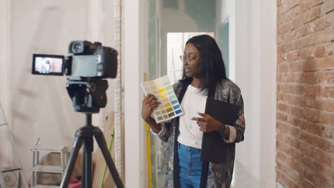 African woman designer choosing paint color for house and recording video for blog. Afro-american decorator blogger filming video on camera having internet blog about renovation