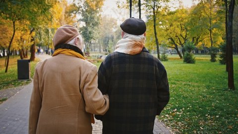 Aged couple, gray-haired wife and husband in elegant outerwear are hugging each other and smiling during romantic walk in the park. Autumn day. Tracking shot, back view. Close up, slow motion