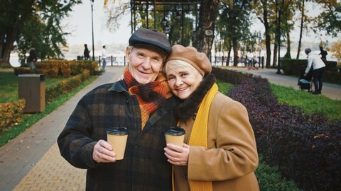 Happy spouses, grandparents holding paper cups of coffee, smiling, hugging and looking at you while posing standing at autumn park during their walk. Close up, copy space. Slow motion, mock up
