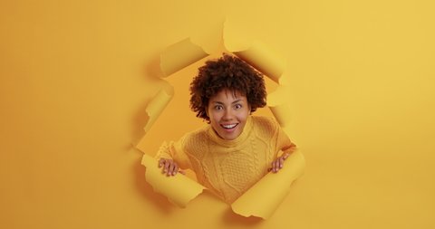 Curious wondered dark skinned woman appears in torn hole has amazed face expression smiles broadly reacts on awesome relevation wears casual jumper poses over yellow background. Wow how great