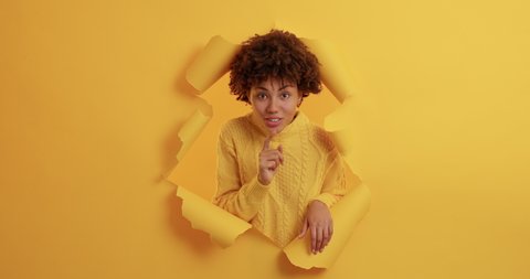 Hey you come here. Pleased cheerful dark skinned woman beckons with inviting gesture invites you to come poses through paper wall of yellow background makes hush sign and looks happily at you