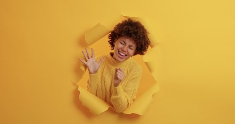 Carefree optimistic dark skinned young woman with curly hair dances happily and sings favorite song keeps hand near mouth as microphone stands in paper hole has happy mood yellow background.