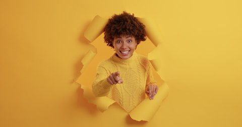 Positive dark skinned young African American woman points directly at camera chooses you picks someone has glad expression dressed in casual yellow jumper stands through torn background. Join me