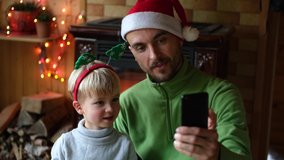 Christmas online video chatting, online greetings. Cheerful father and son using smartphone for virtual call and congratulations family and friends or making selfie photo. New year home celebration.