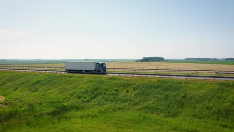 Truck driver with a trailer cargo is driving on intercity speed highway to customer through picturesque agricultural fields on summer sunny day. Delivery and logistics of goods , aerial view motion