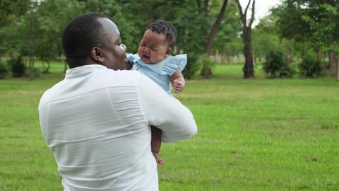 Outdoors, medium shot of happy and kind middle-aged African father cuddle, hold and sing lullaby to calm his crying newborn female colic child at the park