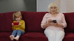 Senior grandmother with child girl granddaughter spending time home together, sitting on sofa in living room, using digital mobile phone. Watching videos, playing games, social network. 6k downscale