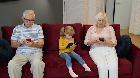 Senior grandparents and granddaughter child girl sit on the couch at home and use mobile phones and digital tablet. Problem in family. Addicted to electronic devices. Look concentrated and antisocial