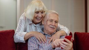 Happy smiling senior couple with mobile phone at home. Cheerful retired husband and wife hugging and laughing using digital smartphone resting on sofa in cozy living room. 6k downscale, slow motion - Powered by Shutterstock - Get 15% off with code: PIKWIZARD15