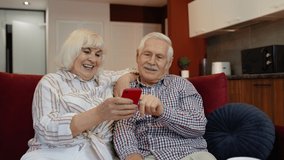 Happy smiling senior couple with mobile phone at home. Cheerful retired husband and wife hugging and laughing using digital smartphone resting on sofa in cozy living room. 6k downscale, slow motion