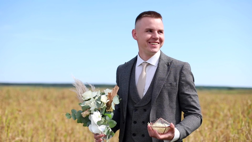 Smiling happy groom holding wedding flowers waiting for bride outside on sunny background. Young family. Lovely couple. Classical grey suit. Celebrating marriage party.