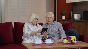Senior old couple grandparents talking and using digital tablet computer at home. Weekend lifestyle, happy elderly grandmother and grandfather doing internet shopping choose sale offers together
