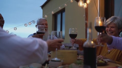 Happy multiracial seniors toasting with red wine glasses together on house patio dinner - Elderly lifestyle people concept 