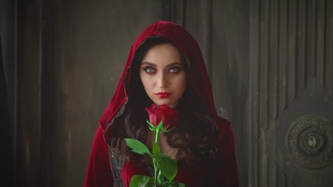 Mysterious beautiful young gothic woman in hood. Closeup girl face red halloween vampire makeup. Sexy fashion velvet vintage red dress. Girl holds rose in hands. Dark room interior medieval castle
