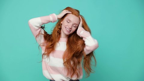 Beautiful cheerful ginger redhead young girl wearing white pink sweater posing isolated on blue turquoise color background in studio. People lifestyle concept. Dancing shaking head with flowing hair