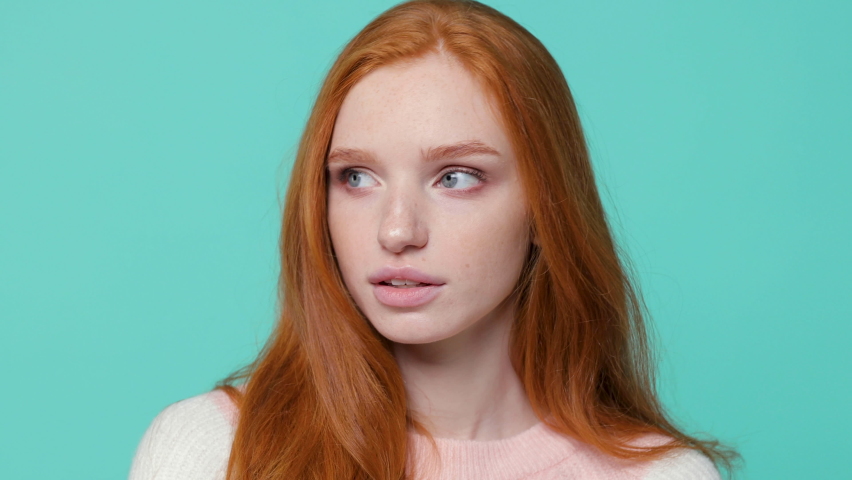 Close up of smiling beautiful ginger redhead young girl wearing white pink sweater posing isolated on blue turquoise color background in studio. People emotions lifestyle concept. Looking camera aside | Shutterstock HD Video #1062391204