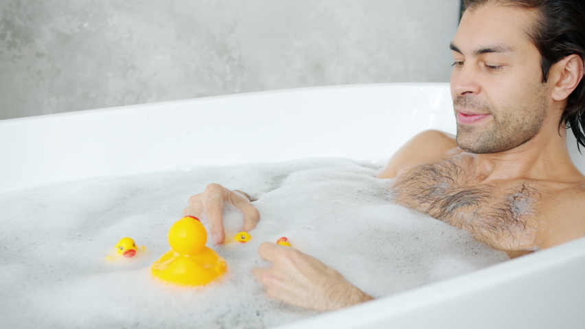 Adult mixed race guy is having with toys playing with rubber ducks in bathtub enjoying carefree activity. Funny games and modern youth concept.