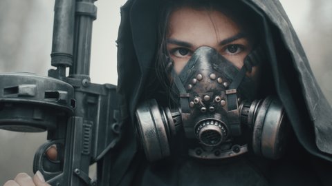 Close-up portrait of woman in grunge clothes hooded mantle and gas mask standing in forest and looking into camera. Dystopian concept, female future survivor. Post apocalyptic world. Cyberpunk soldier