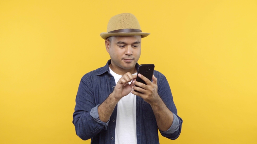 Happy fun asian man in hat hold talk using mobile cell phone chatting just found out great big win news doing ok okay hand gesture isolated on yellow background. Royalty-Free Stock Footage #1062394366