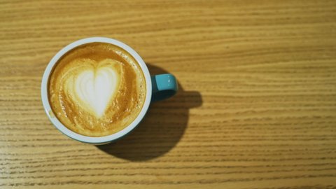 Cup with coffee on wooden background. Latte art of heart design on coffee. Female hands taking cup of hot drink from the wooden surface. Top view.