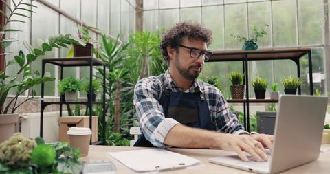 Portrait of Caucasian man entrepreneur in glasses sitting in own flower shop, typing on laptop and writing down in planner. Young male employee in floral store working on computer. Florist concept