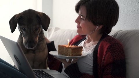 Young woman having video call birthday party at home with her dog - Focus on cake - Social distancing celebration concept 