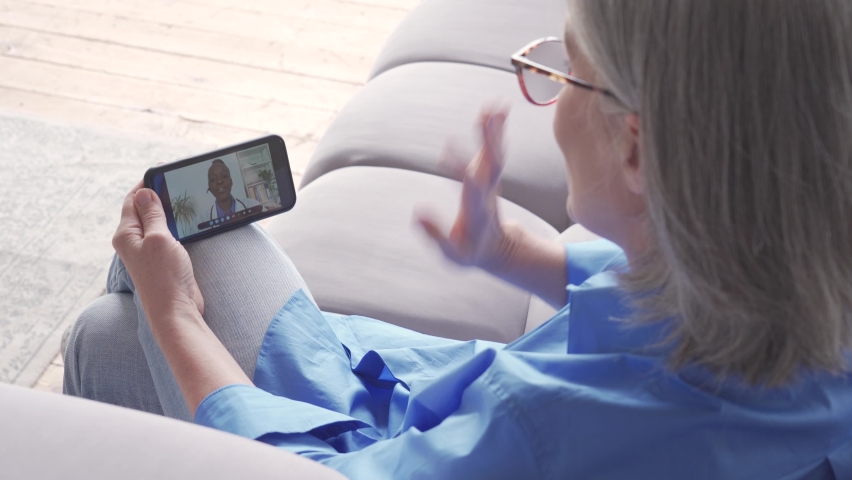 Over shoulder view of older elderly woman patient meeting african virtual doctor using mobile phone at home. Online telemedicine visit. Seniors ehealth, telehealth medical video call consultation.