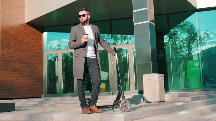 Full shot of successful mixed race businessman in smart clothes and black sunglasses drinking coffee standing near exterior office entrance with e-scooter in daylight