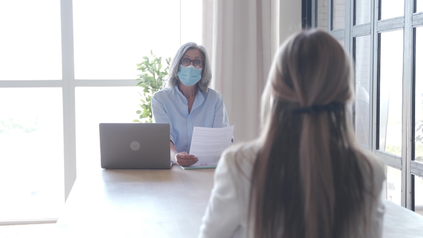 Senior hr recruiter checking female job applicant cv talking at job interview meeting. Old employer wearing face mask reading seeker employment application. Social distance and safety at work concept. Royalty-Free Stock Footage #1062406891